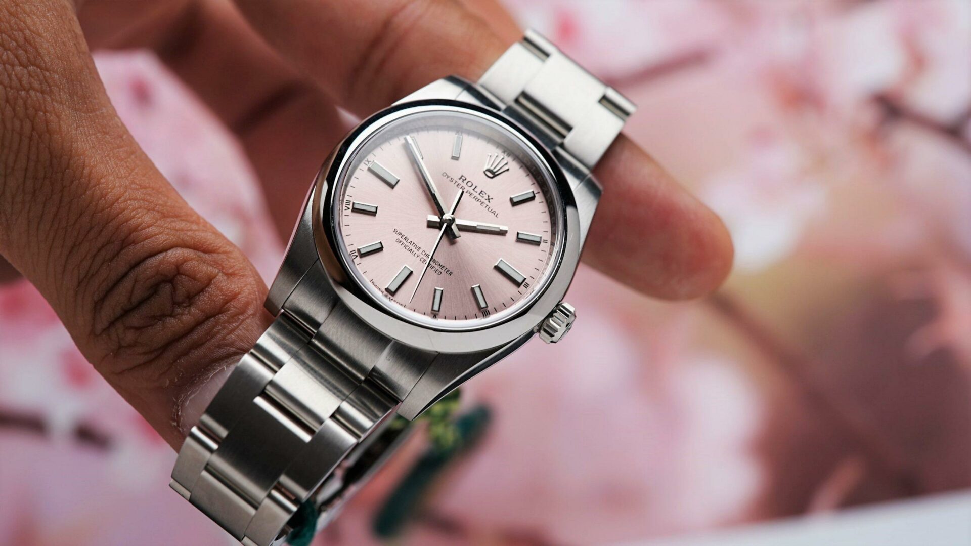 Rolex Oyster Perpetual 34 Pink Dial 2022 held in hand with pink flowers in background.